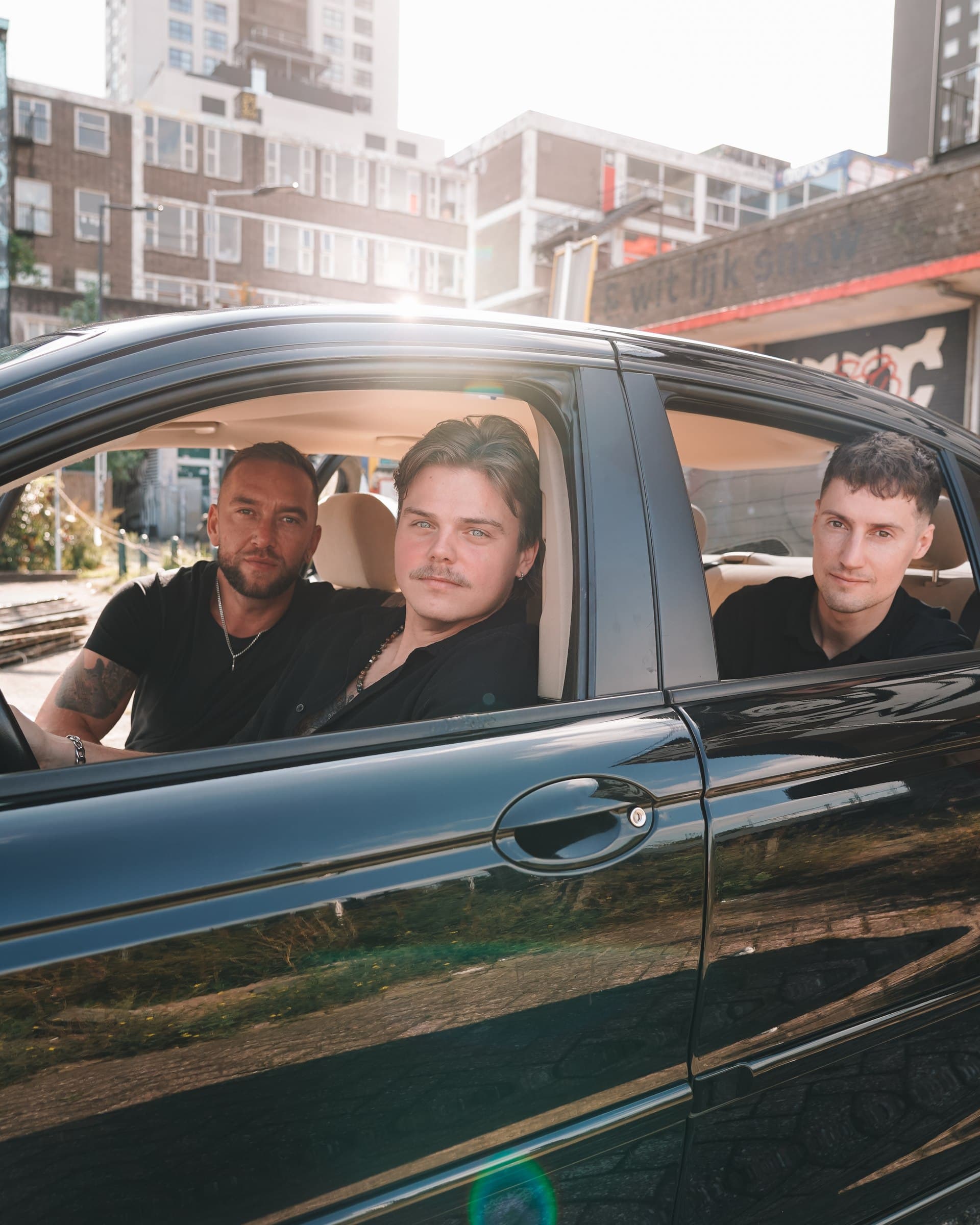 Three Corleone band members looking through the open windows of a black car in sunlight.